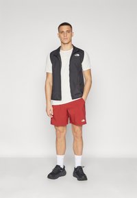 Juodas liemenė virš baltos marškinėlių, raudoni šortai, juodi sportbačiai ir balti kojinės. Ryškus logotipas ant liemenės ir šortų. Paprastas, kasdienis sportinis apranga.