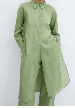 Personne portant une robe-chemise longue vert clair à boutons avec des poches, associée à un pantalon ample assorti, sur un fond uni.