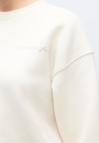 Krēmkrāsas džemperis no mīksta, teksturēta materiāla. Uz krūtīm ir smalks, spīdīgs logo un raglāna piedurknes.