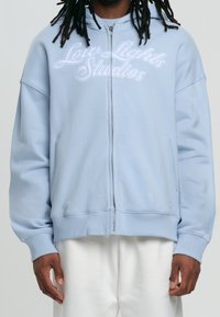 Sudadera con cremallera de color azul claro, con el logo bordado de "Low Lights Studios" en el frente. Incluye capucha, bolsillos laterales y puños ribeteados.