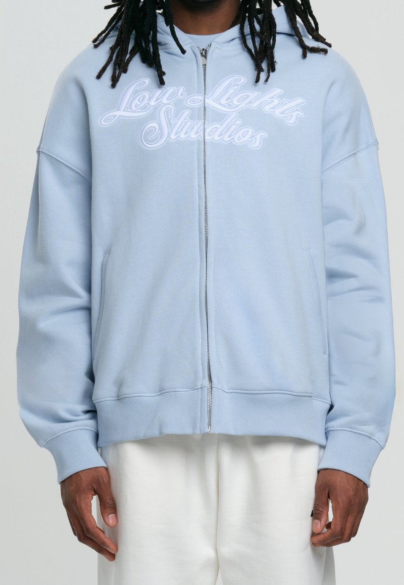 Sudadera con cremallera de color azul claro, con el logo bordado de "Low Lights Studios" en el frente. Incluye capucha, bolsillos laterales y puños ribeteados.