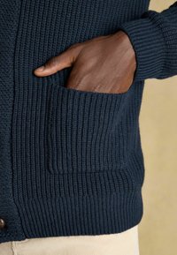 Marineblaue Strickjacke mit gerippter Textur, ausgestattet mit einer Fronttasche und Knopfdetails am Kragen. Nahaufnahme einer Hand in der Tasche.