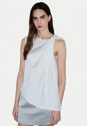Vrouw met lang bruin haar die een mouwloze witte top met fijne strepen en een lichtgrijze satijnen rok draagt, staand tegen een effen witte achtergrond.