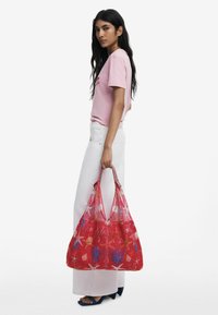 Rote Netz-Tote mit bunten Blumenmustern, mit zwei stabilen Tragegriffen und einem geräumigen Design, die von einer Person in einem pinkfarbenen Shirt gehalten wird.