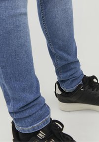 Vaqueros ajustados azules con un ligero desgaste, remangados en los tobillos, combinados con zapatillas negras con suelas y detalles blancos.