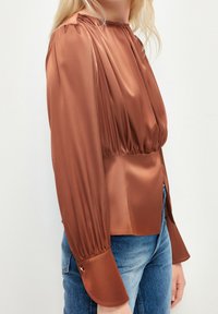 Blouse en satin marron avec des manches plissées, un col froncé et une taille cintrée. Présente des accents de boutons dorés aux poignets.