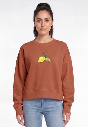Junge Frau mit dunklem Haar im Pferdeschwanz, die einen braunen Pullover mit einem Zitronen- und Zitronenscheibengrafik und hellblaue Jeans trägt.