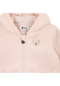 Sweat à capuche en polaire rose clair avec fermeture éclair, orné d'un emblème d'ours sur la poitrine et d'un intérieur doux et texturé. Étiquette de marque visible.