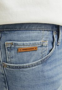 Jeans din denim bleu deschis cu un buzunar din spate cu o etichetă din piele maro inscripționată "SUPERIOR JEANSWEAR" și detalii vizibile de cusătură.