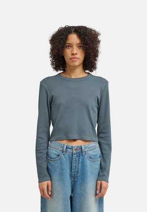 Donna con capelli ricci indossa un crop top blu a maniche lunghe e jeans blu larghi, in piedi rivolta in avanti su uno sfondo chiaro e uniforme.