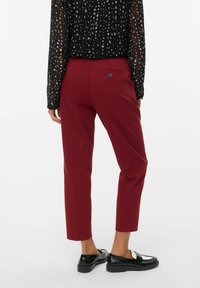 Mujer vestida con pantalones burdeos de largo hasta el tobillo, blusa transparente de patrón en blanco y negro, y mocasines negros, de pie contra un fondo blanco.