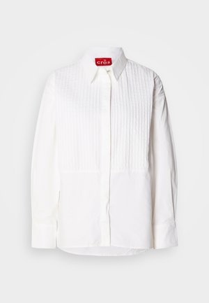 Vit skjorta med en framplacket som har vertikala veck, långa ärmar och en spetsig krage. Etiketten inuti läser "cras." Slät tygtextur.