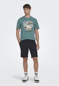 T-shirt turquoise à manches courtes avec un graphisme de plage coloré, assorti à un short noir et des baskets blanches avec des accents noirs.