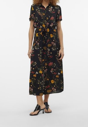 Femme portant une robe midi noire à fleurs, manches courtes et ceinture taille, associée à des sandales à talons ouvertes noires.