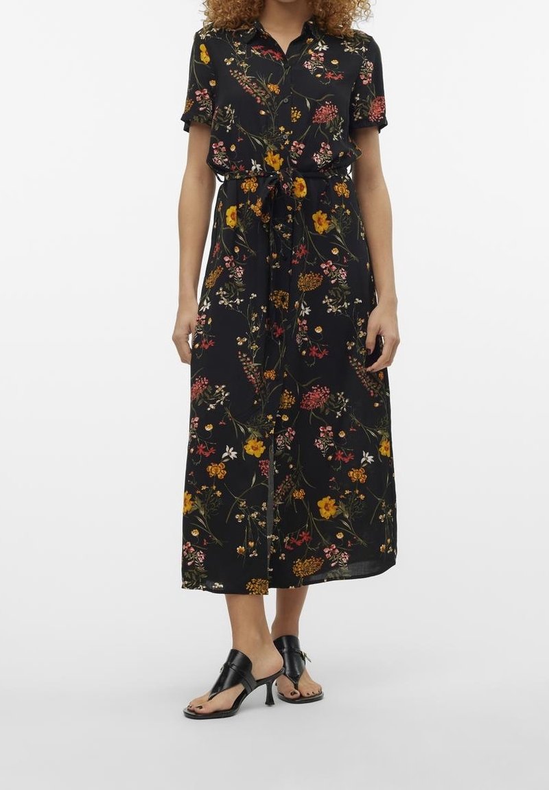 Femme portant une robe midi noire à fleurs, manches courtes et ceinture taille, associée à des sandales à talons ouvertes noires.