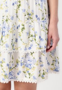Weißes Blumenkleid mit blauen und gelben Blumen, mit gestuftem Design und Spitzenrand am Saum. Leichte Stofftextur.