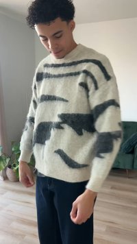 Lichtgrijze gebreide trui met zwarte abstracte patronen, ronde hals en lange mouwen, gecombineerd met een donkere broek. Zacht, getextureerd wollig materiaal.