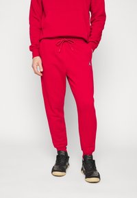 Červená bavlněná mikina s kapucí a odpovídající jogger kalhoty s šňůrkou v pase. Na kalhotách je malá černá značka. Černé sportovní boty s texturovanými akcenty.