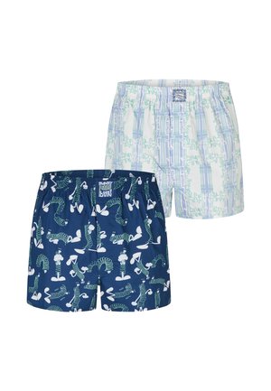 Muskelöser Mann mit kurzen geflochtenen Haaren und Bart, der dunkelblaue Boxershorts mit weißen und grünen abstrakten Mustern trägt und die Hände hinter dem Rücken hält.