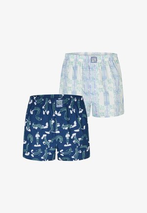 Muskelöser Mann mit kurzen geflochtenen Haaren und Bart, der dunkelblaue Boxershorts mit weißen und grünen abstrakten Mustern trägt und die Hände hinter dem Rücken hält.