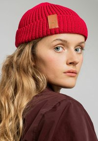 Czerwona wełniana czapka z teksturowanym wzorem i brązową skórzaną naszywką z logo. Czapka ma podwinięty mankiet i dobrze przylega do głowy.