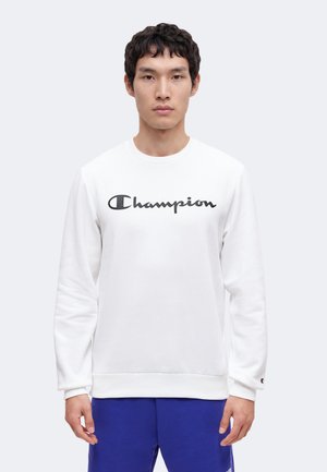 Joven con sudadera blanca con el logo negro de "Champion" y pantalones azules, de pie frente a un fondo claro y liso.