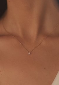 Gouden ketting met een klein, vierkant kristallen hangertje, tegen een gladde huidtextuur, die het licht subtiel weerkaatst.