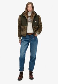 Zielono-brązowa kurtka z futerkiem, zapinana na zamek, w połączeniu z kremowym swetrem z ciemnym wzorem, niebieskimi dżinsami i brązowymi butami na sznurówki.