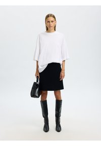 Hvit oversized T-skjorte, svart tettsittende skjørt, knelang svarte skinnstøvler. Modellen holder en svart veske med en sirkulær detalj.