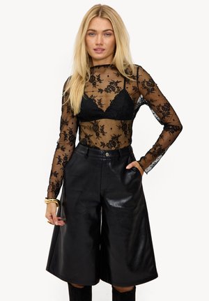 Femme blonde portant un haut noir à manches longues en dentelle florale transparente avec un soutien-gorge noir en dessous et un short large en cuir noir taille haute.