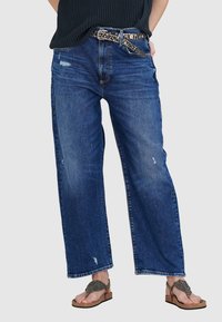 Weite blaue Jeans mit Distressed-Details, ausgestattet mit einem silberbeschlagenen Gürtel und abgetragenen Sandalen, vor einem neutralen Hintergrund.