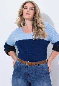 Kvinde med langt blond hår iført en tofarvet blå striktrøje, blå jeans og brunt bælte, stående med hænderne på hoften mod en hvid væg.