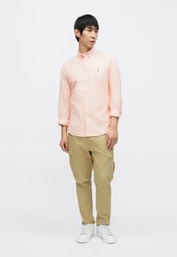 Ανοιχτό ροζ πουκάμισο με κουμπιά και μακριά μανίκια, χακί παντελόνι καμπαρντίνα και λευκά αθλητικά παπούτσια. Διαθέτει μικρό λογότυπο στο πουκάμισο.