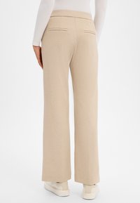 Beige Hosen mit weitem Bein aus strukturiertem Stoff, mit zwei Gesäßtaschen und einem glatten Bund. Kombiniert mit hellen Sneakers.