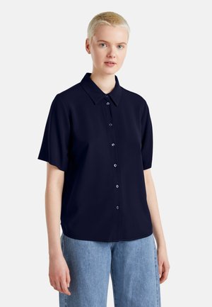Camicia blu navy a maniche corte con chiusura frontale con bottoni, colletto classico e vestibilità morbida, realizzata in tessuto liscio, indossata con denim azzurro.