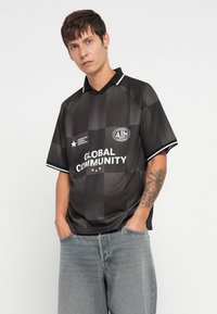 Czarna koszulka piłkarska w kratkę z krótkim rękawem, z kołnierzykiem, białymi akcentami oraz nadrukowanymi logotypami "AIM" i "GLOBAL COMMUNITY". Noszona z jasnymi dżinsami.