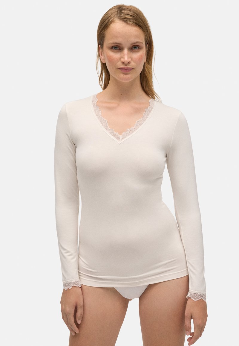 Camiseta de manga larga color crema, hecha de una tela suave con escote en V y detalles de encaje en el cuello y los puños. Diseño simple y ajustado.