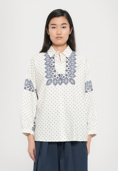 Chemise blanche avec broderie florale bleu marine sur le col et les manches. Dispose d'une patte de boutonnage et de petits motifs étoilés sur l'ensemble.