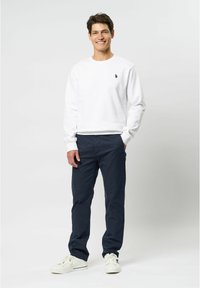 Valkoinen crewneck-huppari, tummansiniset housut ja valkoiset lenkkarit. Hupparissa on pieni logo vasemmalla rinnassa. Yksinkertainen muotoilu, rento istuvuus.