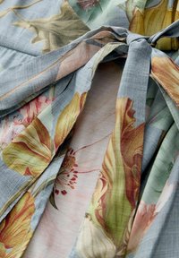 Blau gemustertes Stoff mit floralen Motiven in sanften Pastelltönen, mit einer strukturierten Webart. Bindet sich an der Taille mit einem Riemendetail.