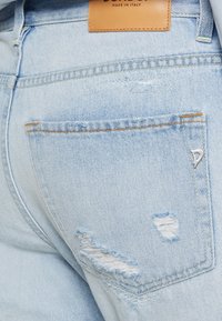 Vaqueros de mezclilla azul claro con un bolsillo trasero desgastado, deshilachados visibles y una etiqueta de cuero beige con la inscripción "Hecho en Italia" en relieve.