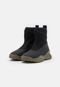 Botas de tobillo negras hechas de un material liso y elástico con una suela beige gruesa. Presentan dos pequeños lazos en la parte superior para facilitar su uso.