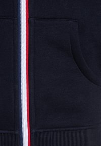 Sudadera con cremallera en color navy que presenta una tela texturizada, dos bolsillos laterales y rayas verticales en rojo, blanco y azul a lo largo de la cremallera.