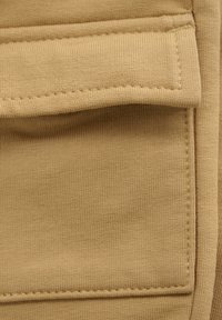 Beige stof met een ribbelstructuur, voorzien van een zak met een patch en zichtbare stikseldetails. Gladde afwerking met een minimalistisch ontwerp.