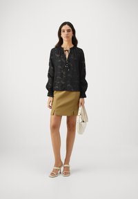 Blusa nera semi-trasparente con scollo rotondo e dettaglio con fiocco, abbinata a una mini gonna khaki con fessure frontali. Sandali bianchi con tacco.