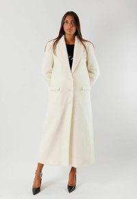 Cappotto lungo in lana color crema con colletto a vista, due tasche anteriori e chiusura a tre bottoni, indossato sopra una maglietta nera.