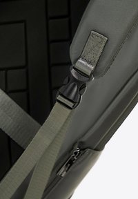 Sac à dos gris avec panneau noir texturé, doté de bretelles réglables, d'une boucle en plastique et d'une poche zippée avec quincaillerie métallique.