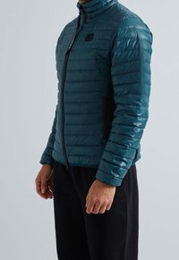Teal pufferjacka med hög krage, dragkedjestängning och slim fit. Innehåller horisontell sömnad och diskret logotyp på bröstet. Svarta detaljer.
