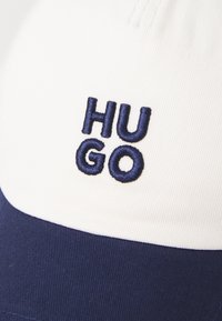 Bílá a námořnická baseballová kšiltovka vyrobená z bavlny, s vyšitým logem "HUGO" v tmavě modré barvě na přední straně a zakřiveným námořnickým kšiltem.