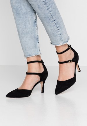 Czarne zamszowe buty na wysokim obcasie z ostrym czubkiem, z paskami wokół kostki i jasnym obcasem. Noszone z postrzępionymi niebieskimi dżinsami.